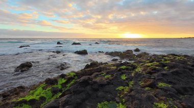 Göz kamaştırıcı bir gün batımı dalgalı yeşil yosunlarla bezenmiş kayalık sahili aydınlatıyor ve gerçekten sakin bir kıyı manzarası yaratıyor.