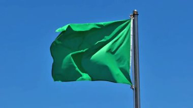 Parlak yeşil bir bayrak zarif bir şekilde mavi gökyüzüne doğru dalgalanıyor ve büyüleyici bir görsel etki yaratıyor.