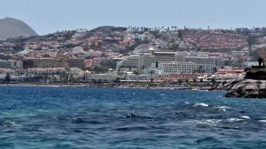 Parlak mavi denizin yanında güzel, canlı bir şehir manzarası gösteren nefes kesici bir sahil manzarası.