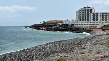 Huzurlu ve güzel bir kıyı manzarası deniz kenarında modern bir otelin yanında kayalık bir plaj sergiliyor.