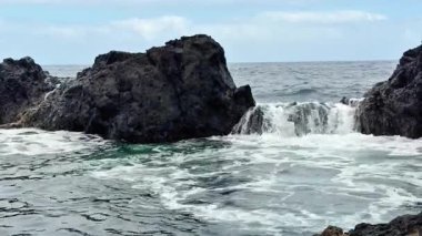 Dinamik okyanus dalgaları, engin mavi gökyüzünün altındaki kayalıklara çarpar ve çarpıcı bir manzara yaratır.
