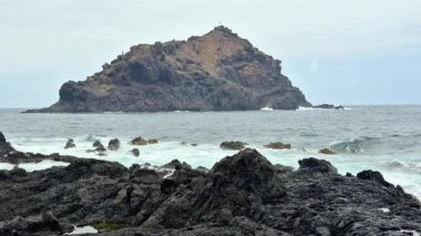 Nefes kesici bir kayalık kıyı şeridi, kasvetli gökyüzünün ve çarpan dalgaların altında görünen uzak bir adaya sahiptir.
