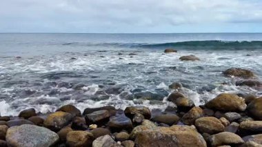 Huzurlu ve sakin bir okyanus manzarası engebeli kayaları ve bulutlu gökyüzünün altında dalgaları sergiliyor.