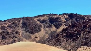 Uçsuz bucaksız, kurak bir çölde, dalgalı mavi gökyüzünün altında sıcak, yumuşak kum tepeleri olan sarp ve kayalık tepeler yükselir.