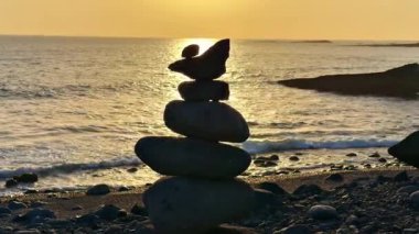 Güzelce dingin bir sahil sahnesi dikkatlice dengelenmiş taşları sergiliyor nefes kesici bir günbatımı arka planına karşı.