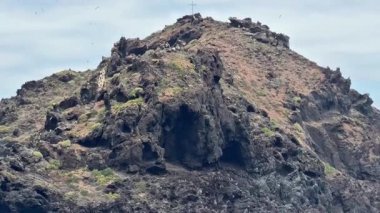 Çarpıcı bir manzara, zirvesinde bir haç bulunan volkanik bir kaya oluşumunu betimler.