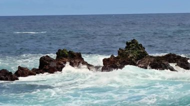 Okyanus dalgaları engebeli kayalara karşı zarif bir şekilde dans ediyor, doğanın muazzam gücünü güzel bir şekilde sergiliyor.