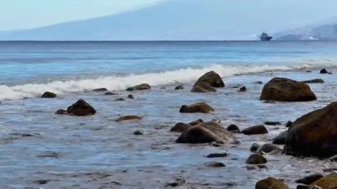 Sakin ve sakin bir sahil şeridi. Hafif dalgalar, kayalara çarpan kayalar ve sakin sularda seyreden uzak bir gemi.