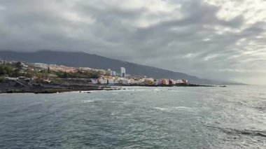 Dramatik ve karamsar gökyüzüne karşı inşa edilmiş canlı ve renkli binaları gösteren nefes kesici bir kıyı manzarası.