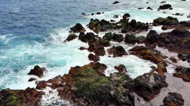 Dalgaların kıyıya çarptığı kayalık bir kıyı şeridinin çarpıcı hava manzarası canlı bir deniz manzarası yaratıyor.