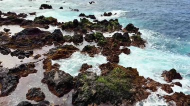 Çarpıcı manzara okyanus dalgalarının engebeli bir kıyıya çarptığını gösteriyor. Canlı, yemyeşil yosunlarla süslenmiş.