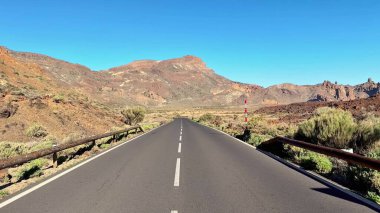 Göz kamaştırıcı bir manzara, açık, parlak mavi bir gökyüzünün altında, pitoresk dağların arasından dolambaçlı bir yol sunar.