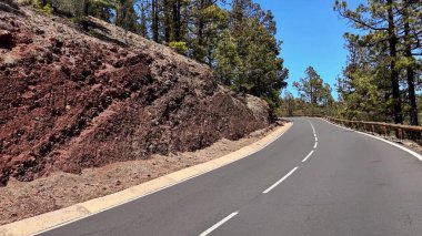 Güzel, dolambaçlı bir yol yeşil ağaçların arasından geçiyor ve açık mavi gökyüzünün altında kayalara çarpıyor.