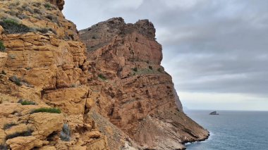 Göz kamaştırıcı ve güzel manzaralar bulutlu bir gökyüzünün altındaki engebeli kıyı şeridini çevreleyen kayalık uçurumları gösteriyor.