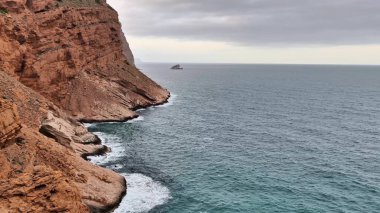 Dramatik kıyı manzarası kayalık uçurumlar ve bulutlu bir gökyüzünün altında kırılan sakin okyanus dalgalarını içerir.