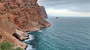 Çarpıcı bir kıyı manzarası bulutlu gökyüzünün altında kızıl kayalıkları, yumuşak dalgaları ve sakin suları gösteriyor.