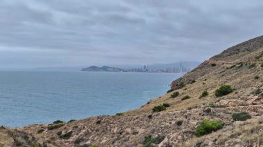 Çarpıcı bir sahil manzarası engebeli tepeleri sergiliyor uzak bir şehrin silueti ile buluşuyor, bulutlu gökyüzünün altında.