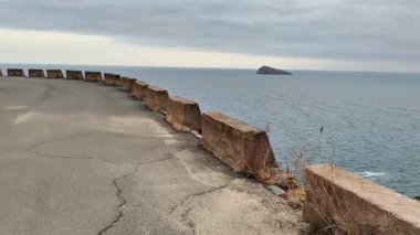 Sakin ve sakin bir kıyı manzarası bulutlu bir gökyüzünün altındaki uzak bir adaya doğru kıvrımlı bir yola sahip.