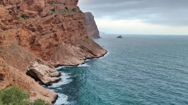Çarpıcı bir kıyı manzarası bulutlu gökyüzünün altında kızıl kayalıkları, yumuşak dalgaları ve sakin suları gösteriyor.