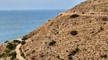 Bu çarpıcı hava görüntüleri, okyanus kıyısındaki yeşil bir tepeyi saran toprak bir yolu gösteriyor. Engebeli araziyi sakin sularla harmanlıyor, keşifleri teşvik ediyor.
