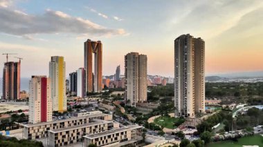 Bir hava manzarası, kentsel hareketliliğin ortasında çarpıcı bir gökdelen silueti sergiliyor. Manikürlü bir golf sahası, modern şehir yaşamlarını ihtişam ve sükuneti yansıtan bu canlı manzaraya zıt düşüyor.