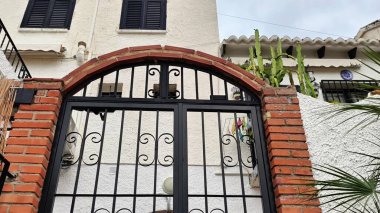 Video, tuğla bir duvar, beyaz bir bina ve yakınlardaki tuğla bir yapı tarafından çerçevelenmiş işlenmiş demir kapılı büyüleyici bir ortam sergiliyor.