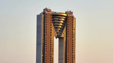 Bu video, benzersiz bir tasarımı ve merkezi deliği olan, açık mavi gökyüzüne karşı kurulmuş, modern şehir yaşamını gözler önüne seren uzun bir gökdeleni içerir.