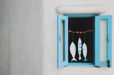 Beyaz alçı duvarda asılı pastel mavi pencerenin yatay görüntüsü. Önünde mavi ve beyaz renkli şirin bir balık gezgini var. Metin için alanı olan Akdeniz kıyı dekoru.