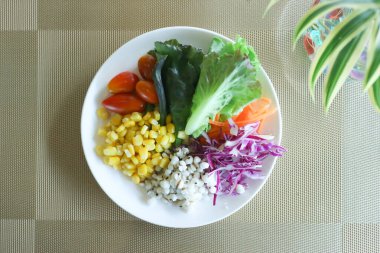 Domates, sarı mısır, koix tohumu, mor lahana, havuç ve yeşil koza içeren altı çeşit sebzeden oluşan güzel bir salatanın üst görüntüsü. Çevre dostu yaşamı sembolize eden dekoratif bir bitkiyle altın bir yerde sunuluyor..