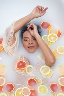 Limon ve portakal dilimleriyle çevrili süt banyosunda güzel bir kadın. Kolları havada.