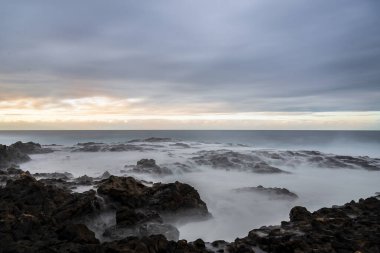 Bulutlu bir gökyüzünün altında vahşi, volkanik bir kıyı şeridine dramatik ve atmosferik bir şekilde uzun süre maruz kalmak. Sisli, akan su güçlü ve ruhani bir manzara yaratır..