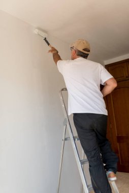 Ressam, sağlam metal bir merdiven üzerinde denge kurarken duvara taze beyaz boya uyguluyor ve evin içini büyütüyor.