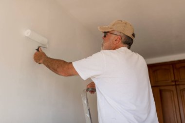 Son sınıf boyacı çalışıyor, duvarı beyaza boyuyor boya rulosu kullanıyor, merdivenin üstünde duruyor.