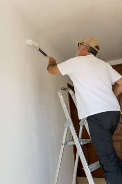 Son sınıf boyacısı çalışıyor, beyaz bir duvar boyuyor, merdivenin üstünde duran boya silindiri ile.