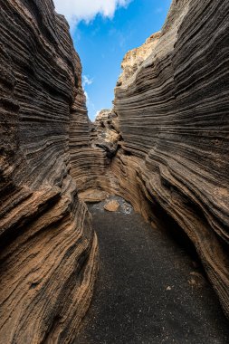 Dar bir kanyonun manzarası, katmanlı volkanik kaya oluşumları berrak mavi bir gökyüzünün altında güneş ışığıyla aydınlatılıyor. Peyzaj ve doğa kavramı