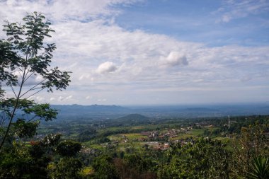 Geniş panoramik bir manzara, küçük evler ve pirinç tarlalarıyla kaplı, mavi gökyüzünün altında, dağınık bulutlar, yüksek bir noktadan ön plandaki ağaçların arasından alındı..