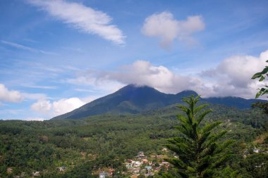 Zirvelerin etrafını saran bulutlarla dolu canlı bir dağ manzarası, parlak mavi gökyüzünün altındaki renkli köy evleriyle benekli yemyeşil bir vadiye bakıyor..