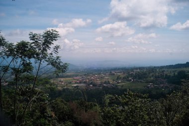 Yeşil tepeler ve ağaçlarla çevrili, dağınık mavi bulutlarla kaplı kırsal bir vadide gündüz manzarası..