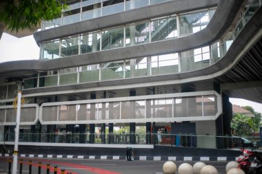 Parlak cam ve metal cepheli modern bir toplu taşıma istasyonu, çok katlı ve yürüyen merdivenli. Ön planda kalın çizgiler, kırmızı boyalı yaya geçidi ve yol boyunca dekoratif beton küreler yer alıyor..