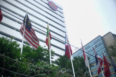 ASEAN üyesi ülkelerden gelen bir dizi ulusal bayrak, ASEAN logosunun bulunduğu modern bir binanın önünde dalgalanıyor. Sahnede tropikal yeşillik ve ön planda tel örgü yer alıyor..