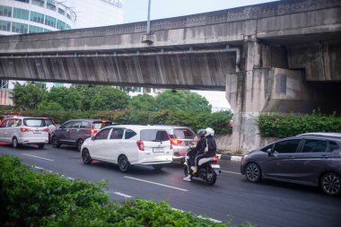 Yoğun trafiği olan hareketli bir şehir caddesi. İçinde arabalar ve güvenlik ekipmanları olan bir motosikletçi var. Sahnede yol kenarında beton bir üst geçit ve yeşil bitki örtüsü bulunuyor..