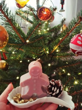 Arka planda süslü bir Noel ağacı olan seramik tabakta zencefilli adam mumu ve çam kozalağı tutan bir elin yakın çekimi. Sıcak ve şenlikli tatil atmosferi.