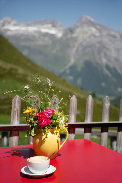 Солнечная летняя сцена с чашкой кофе и желтым кувшином цветов на красном столе, установленном на живописном альпийском фоне. Концепция отдыха, отдыха и путешествий.