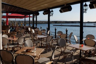 Deniz kenarında boş bir kafeterya terası. Hasır sandalyeler ve masalar hazır güneşli bir sabahta müşterileri bekliyorlar. Sakin su manzaralı bir Akdeniz tatil köyünde sakin bir manzara.