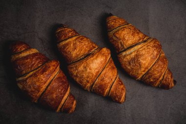 Three freshly baked croissants sit on a dark grey countertop, ready to be devoured. Their golden-brown crust glistens under the soft light, inviting a bite. The rich aroma of buttery pastry fills the air, promising a delicious start to the day. clipart