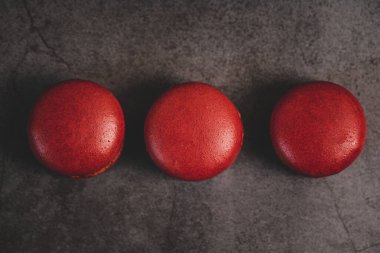 Üç mükemmel yuvarlak kırmızı badem kurabiyesi koyu gri bir levha yüzeyinde sıralanarak görsel olarak çekici ve simetrik bir bileşim oluşturur. Makaronların pürüzsüz, parlak yüzeyi ışığı yansıtır ve canlı kırmızı renklerini güçlendirir..