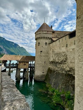 İsviçre 'nin Montreux kenti yakınlarındaki Cenevre Gölü kıyıları boyunca inşa edilmiş tarihi taş kale, turkuaz su üzerinde ahşap bir köprü ve arkaplanda parçalı bulutlu bir gökyüzünün altında manzaralı alp dağları bulunuyor..