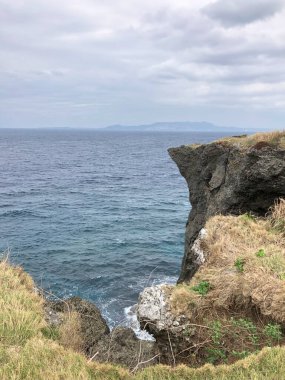 Dik, çimenli bir uçurum Okinawas kıyı şeridi boyunca okyanusa dalıyor. Dalgalar alttan vuruyor ve bulutlu bir gökyüzünün altında uzak bir ada görülebiliyor..