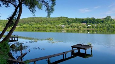 Yumuşak mavi bir gökyüzünün altında, kırsal ahşap bir rıhtım ve yemyeşil ağaçlar tarafından çerçevelenmiş sessiz bir göl manzarası belirir..