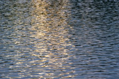 Işıldayan mavi suyun azgın güzelliği güneş ışığı altında hafifçe dalgalanırken altın renkler yüzeyi boyunca dans ediyor.